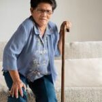 An elderly woman struggles to stand up from sitting on a couch using a cane before calling a Medicare Lawyer Des Moines, IA